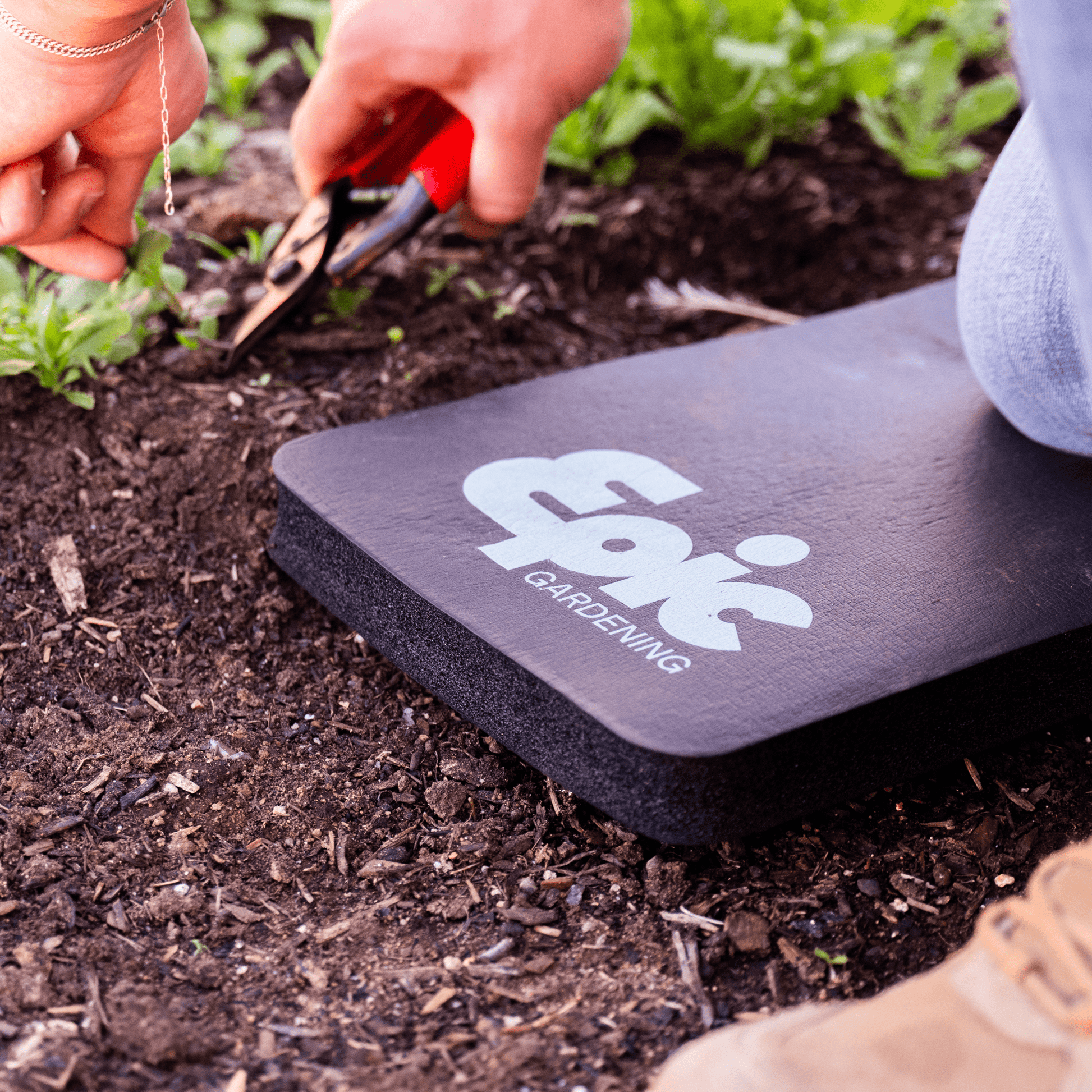Gardener kneeling on a black kneepad for gardening, holding pruning shears. The brand name Epic Gardening appears on the pad.