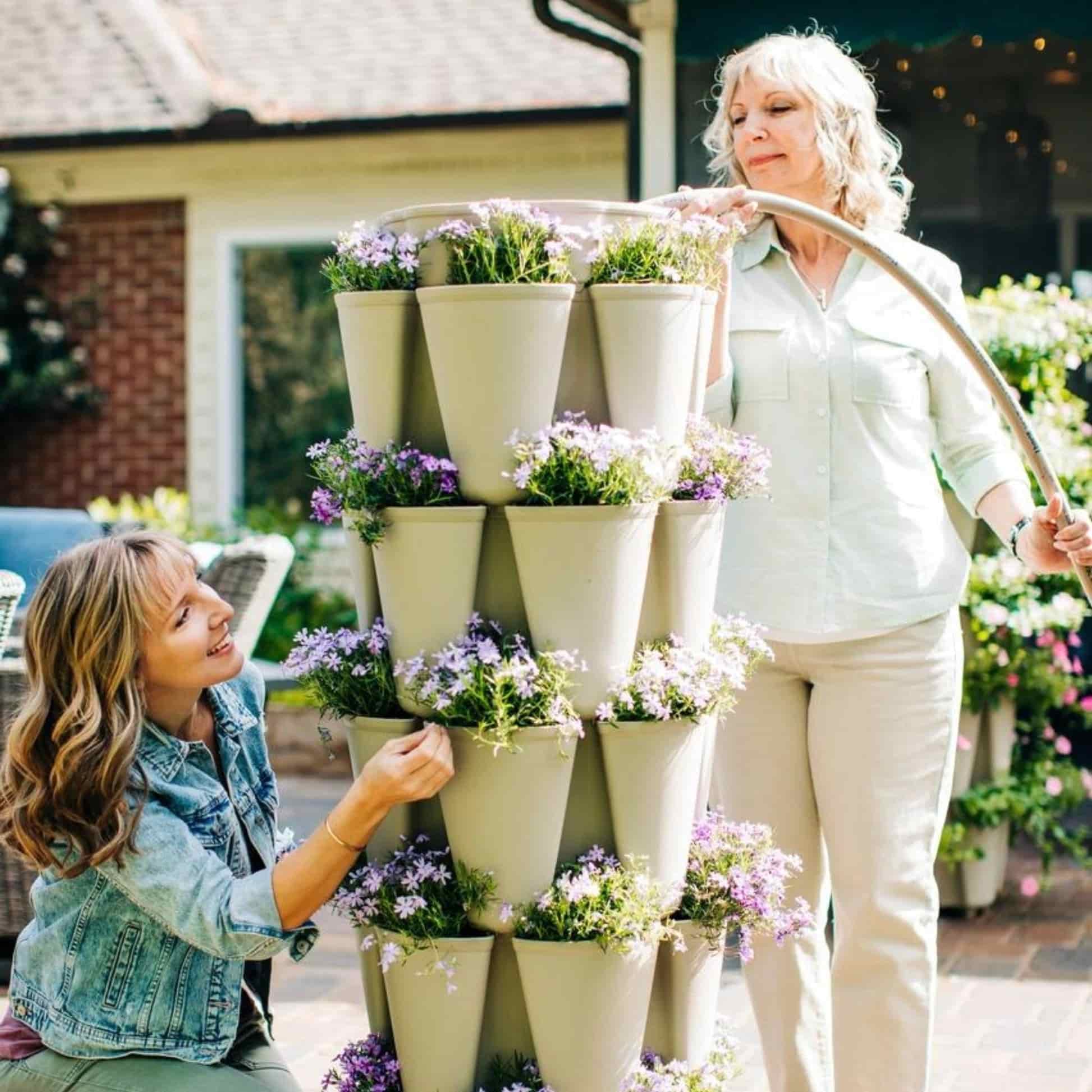 Two women tend a stacked, five-tier stone-coloured GreenStalk vertical planter overflowing with purple and white flowers, one kneeling to touch the blooms while the other waters it with a hose on a sunny patio.